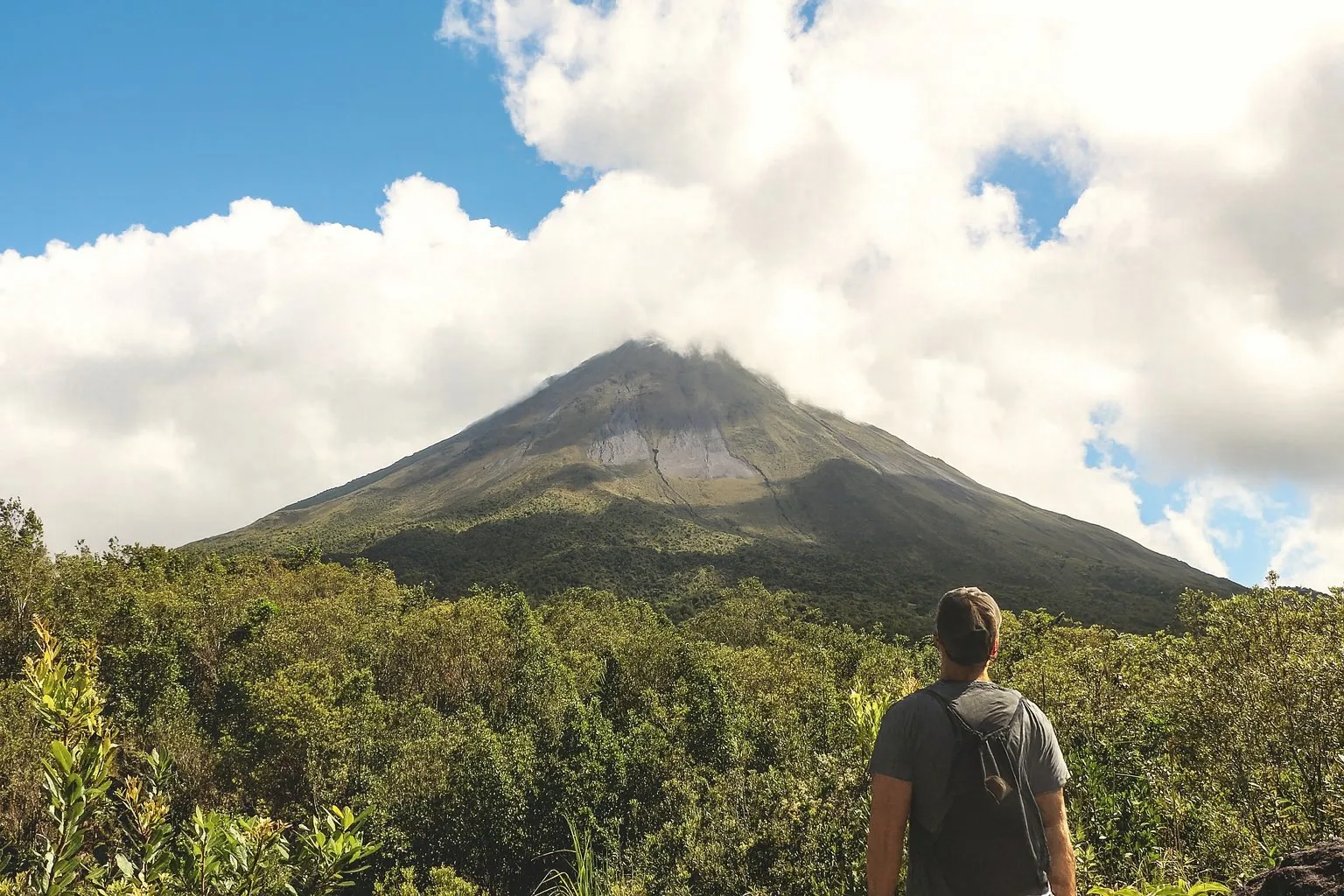 A hiker looking up at Arenal Volcano under a morning cloud cap, near La Fortuna