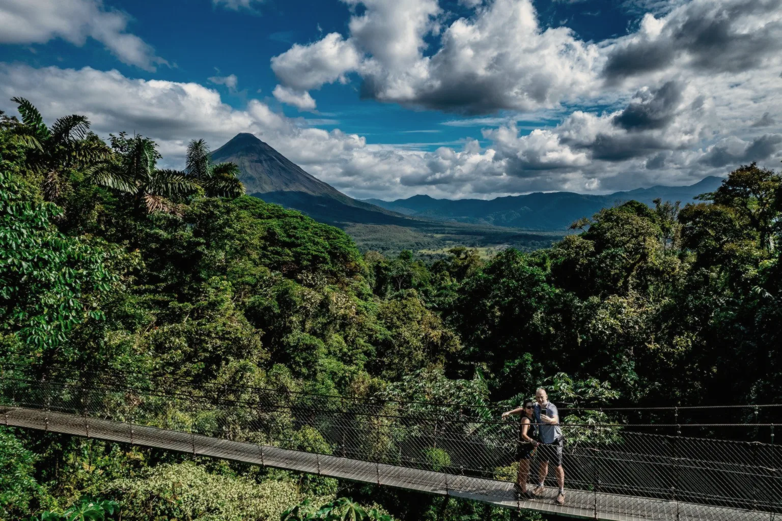 Two travelers on a hanging bridge above the rainforest canopy with Arenal Volcano behind, near La Fortuna
